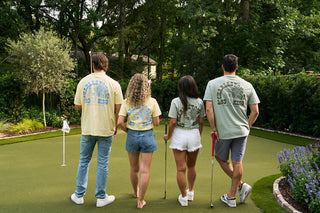 Four people on a mini golf course wear pastel SDS 'King Street' Flagship Tees with “Charleston 1938” on the back. Surrounded by greenery, they enjoy a stylish, comfortable day thanks to Sewing Down South’s signature shirts.