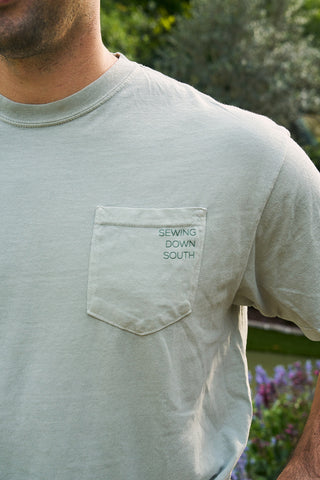 Wearing an SDS 'King Street' Flagship Tee in light gray heavyweight cotton, featuring a green "SEWING DOWN SOUTH" chest pocket embroidery, a person stands amid lush greenery and blurred flowers, capturing a relaxed, summery vibe.