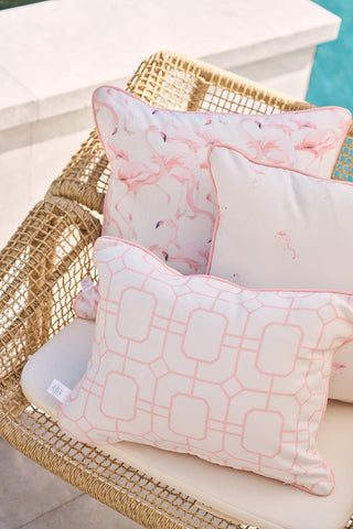 Three decorative pillows made from SDS Shield® Outdoor Fabric are on a beige wicker chair by the pool. The front shows a pink geometric pattern, the middle has playful pink flamingos (the Flamingo Trio), and the back is plain white with subtle flamingo accents.
