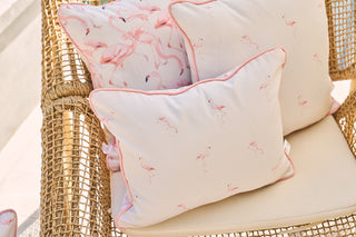A wicker chair is adorned with two Flamingo Scatter Outdoor Pillows, showcasing pink flamingo designs. In a sunny outdoor setting, the intricate woven texture and delicate SDS Shield® Outdoor Fabric offer vibrant durability.