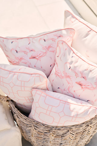A woven basket filled with outdoor pillows, including the Flamingo Trio with Flamingo Cluster, showcases vibrant pink flamingos on a white background while others display a light pink geometric design. This colorful collection rests against a bright surface.