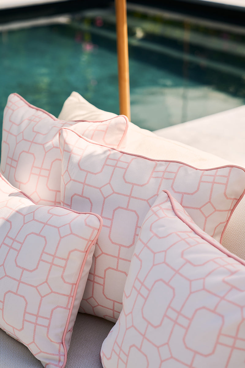 Four Pink Lattice Outdoor Pillows crafted with SDS Shield® Outdoor Fabric adorn a light sofa by the aqua pool, enhancing charm and durability. A wooden umbrella pole complements the serene setting.