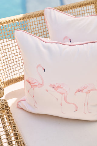 Two Flamingo Trio with Flamingo Cluster outdoor pillows, made from durable SDS Shield® Outdoor Fabric, are placed on a wicker chair. The backdrop features a blurred poolside area, creating a sunny and inviting outdoor vibe.