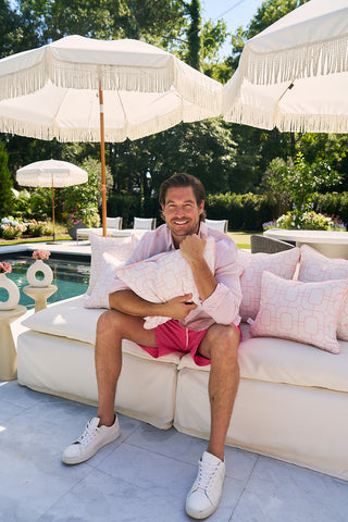 A person with dark hair and a beard smiles while sitting on an outdoor couch, holding a Flamingo with Pink Lattice Lumbar Outdoor Pillow under a fringed umbrella by the pool. Theyre wearing a light pink shirt, bright pink shorts, and white sneakers, surrounded by trees in the background.
