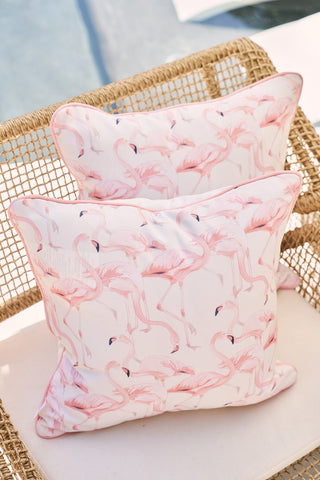 Two decorative outdoor pillows with the Flamingo Trio pattern on a wicker chair feature SDS Shield® Outdoor Fabric against a white backdrop. The scene is outdoors, with faint shadows and water visible in the background.