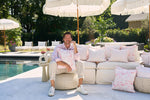 A person in a light pink shirt and beige pants sits on a woven pouf, surrounded by Flamingo Trio with Flamingo Cluster Outdoor Pillows on a white sofa. Theyre outdoors by the pool, with white umbrellas and lush greenery, enjoying the comfort of durable SDS Shield® Outdoor Fabric.