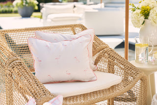 A wicker chair featuring a Flamingo with Pink Lattice Lumbar Outdoor Pillow and white cushion adds a pop to the patio. Nearby, a small table holds lemon water and white flowers, while extra seating with outdoor fabric is visible against the blurred backdrop of a garden view.