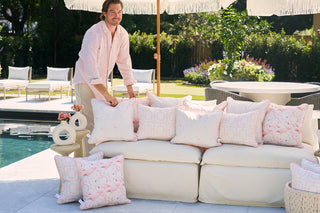 In a garden with lawn chairs, a person in a light pink shirt arranges Flamingo Scatter Outdoor Pillows on an outdoor sofa by the pool under a sunny sky, complemented by a large umbrella and enhanced by durable SDS Shield® Outdoor Fabric.