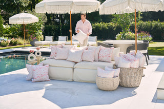 A man smiles by the poolside, holding a Flamingo with Pink Lattice Lumbar Outdoor Pillow. He stands beside a large sofa adorned with white and pink lattice pillows under shaded umbrellas. Lush greenery and extra seating in the background enhance the relaxed, inviting vibe.
