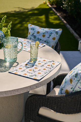 Outdoor table setting with two wicker chairs featuring floral cushions and Blossoming Peonies Oversized Outdoor Pillow. A round table adorned with floral placemats, a textured glass pitcher, and a lemon-infused water glass completes the scene, sunlit shadows and greenery background enhancing the ambiance.