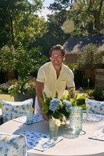 A person in a yellow shirt is cheerfully setting a round outdoor table with floral cushions and placemats. The table features a vase with blue hydrangeas and two glasses, while sunlight highlights the Blossoming Peonies Oversized Outdoor Pillow in this garden setting.