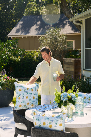 A man in a light yellow shirt stands by an outdoor table featuring a Blossoming Peonies Oversized Outdoor Pillow and a drink pitcher. He holds a beverage, grinning in the sunny garden, with blooming peonies and trees framing the house in the background.