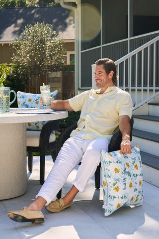 A man in a yellow shirt and white pants sits on a patio chair, drink in hand, smiling and relaxed. Nearby is the Blossoming Peonies Oversized Outdoor Pillow. The scene is outdoor with a table, stairs, and house in the backdrop.
