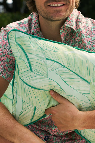 Dressed in a floral shirt, someone holds a Banana Leaves with Green Lattice Lumbar Outdoor Pillow. Their lower face peeks through, set against a beautifully blurred green backdrop, evoking serene elegance.