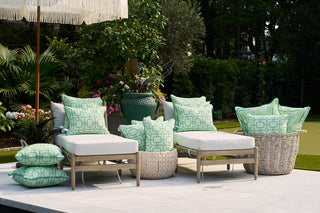 An outdoor patio scene features two cushioned lounge chairs with Green Lattice Outdoor Pillows in mint green geometric patterns, accessorized by a side table and greenery on a light stone patio.