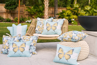 The outdoor seating area showcases a woven chair and crocheted ottoman, each adorned with the Butterfly Scatter Outdoor Pillow in blue and yellow. Lush greenery and a wooden fence create a serene backdrop for this inviting space.