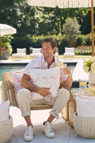 A smiling man sits on a wicker chair by the pool, holding a Flamingo Trio with Flamingo Cluster Outdoor Pillow while enjoying a drink with lemon. Dressed in a light floral shirt and beige pants, hes surrounded by vibrant flowers, cushions, and an umbrella under the sunny sky.