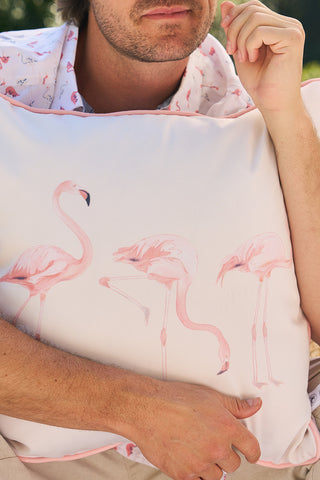A person with a slight beard holds the Flamingo Trio with Flamingo Cluster Outdoor Pillow, made from durable SDS Shield® Outdoor Fabric. They sport a flamingo-patterned shirt in lush greenery, enjoying the vibrant outdoor setting.