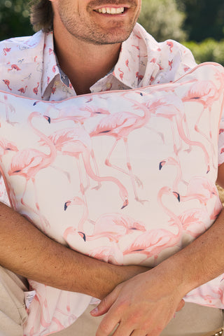 A person sits outdoors, smiling and clutching a Flamingo Trio with Flamingo Cluster Outdoor Pillow while wearing a matching shirt. The blurred green background suggests a sunny day, ideal for enjoying SDS Shield® Outdoor Fabric products.