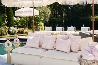 The poolside seating area features cream cushions with geometric designs and a Pink Lattice Outdoor Pillow, all enhanced by SDS Shield® Outdoor Fabric. Umbrellas provide shade, while small tables hold vases of pink flowers, complemented by lounge chairs and lush greenery.
