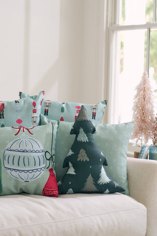 A cozy sofa features the Festive Tree Shape Pre-Filled Pillow among other festive throw pillows. Nearby, a small pink Christmas tree and a basket sit on a table by a sunlit window.