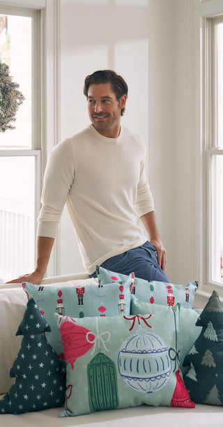 A man in a cream sweater and jeans sits on a white sofa decorated with holiday pillows, including the Festive Ornament Pre-Filled Lumbar Pillow, in a bright room with large windows.