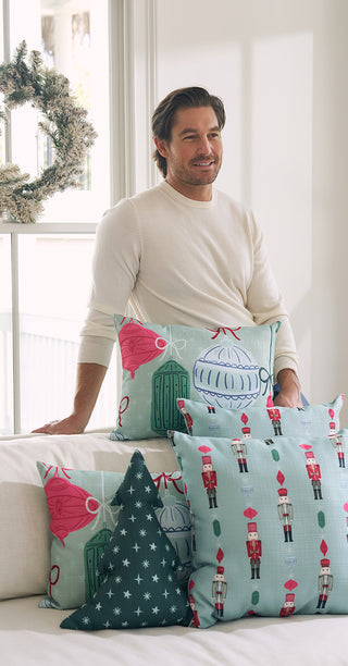 A man in a cream sweater sits on a white couch with the Festive Holiday Pillow Bundle: Get 1 of Each!—pillows featuring nutcrackers, ornaments, and stars—in a bright room with a window and wreath.