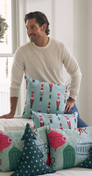 A man in a cream sweater leans on a couch with Festive Nutcracker Pre-Filled Pillows, which display blue, pink, and green nutcracker and ornament designs. Natural light streams in through the window behind him.