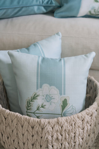 Two light blue decorative pillows with floral and leaf patterns, possibly from Sewing Down South®, sit in a beige knitted basket. In the background, part of a light-colored couch and another pillow hint at Victorian Floral Stripe elegance from the Bee & Florals Collection.
