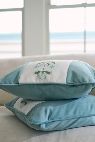 Two Victorian Floral Stripe Pillows from the Bee & Florals Collection are stacked on a light-colored couch. A window in the background reveals a blurred view of the ocean and sky, enhancing the serene ambiance.