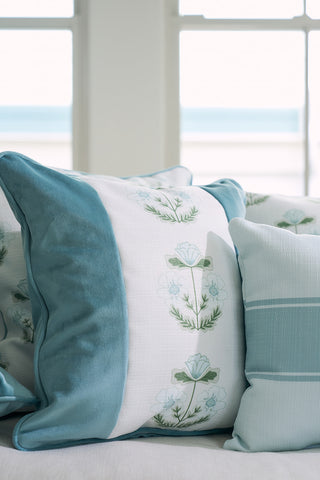 A cozy bed features a Victorian Floral Stripe Pillow from the Bee & Florals Collection, adorned with elegant green leaves and blue flowers on plush teal velvet. Pastel pillows complement this Sewing Down South® design, while a window in the background offers a serene view.