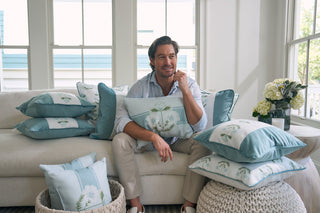 A person sits on a light-colored couch holding a decorative pillow from the Bee & Florals Collection: Victorian Floral Stripe Pillow, surrounded by similar designs. Baskets and a vase of flowers are nearby, while large windows in the background let natural light pour in.