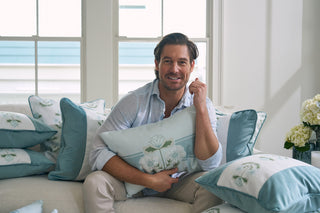 A person with short dark hair sits on a couch, surrounded by blue and Bee & Florals Collection: Victorian Floral Stripe Lumbar Pillows. They smile in a light blue shirt and beige pants as sunlight streams through large windows, highlighting the cozy setting with USA-made Sewing Down South® designs.
