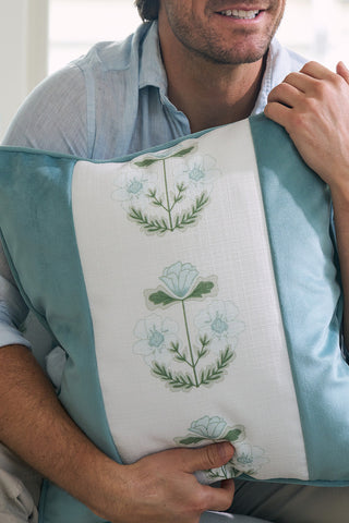 A person in a light blue shirt sits holding a Victorian Floral Stripe Pillow from the Sewing Down South® Bee & Florals Collection. The pillow features light blue and green floral embroidery on a white stripe with solid blue borders, with the persons face partially visible.