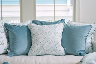 A collection of decorative pillows from Sewing Down South adorns the sofa in a bright room with large windows, showcasing soft blue and white designs. The Bee & Florals Collection: Victorian Tile Pillow adds geometric and floral motifs, accentuated by a teal velvet welt for extra elegance.