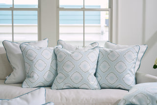 A cozy sofa is embellished with Victorian Tile Pillows from the Bee & Florals Collection, showcasing a pale blue and white geometric design with teal velvet borders. Positioned in a bright room with large windows, it beautifully welcomes an abundance of natural light.