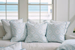 A cozy sofa is embellished with Victorian Tile Pillows from the Bee & Florals Collection, showcasing a pale blue and white geometric design with teal velvet borders. Positioned in a bright room with large windows, it beautifully welcomes an abundance of natural light.