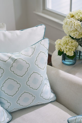 A cream sofa with patterned blue and white cushions, featuring a Victorian Tile Pillow from the Bee & Florals Collection, sits next to a round white table with a bouquet of white hydrangeas in a blue vase, bathed in soft natural light.