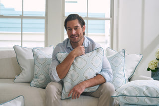 A man relaxes on a couch with a Bee & Florals Collection: Victorian Tile Pillow amid similar cushions. Smiling in a light blue shirt and beige pants, the sunlit room showcases his fondness for Sewing Down South decor.