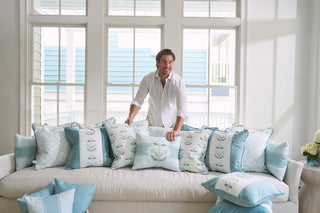 A person in a white shirt stands behind a couch with Bee & Florals Collection: Victorian Floral Stripe Lumbar Pillows from Sewing Down South®, featuring light blue floral patterns. Large windows reveal an exterior view with blue siding, creating a classic yet modern ambiance.