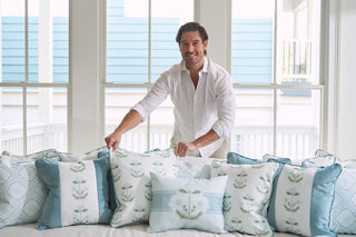 A man in a white shirt smiles as he arranges Bee & Florals Collection: Victorian Floral in Blue pillows featuring blue, white, and Victorian floral patterns on the bed. Large windows in the background fill the room with natural light.