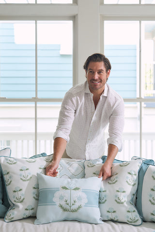 A person in a white shirt is arranging decorative pillows, including the Bee & Florals Collection: Victorian Floral Stripe Lumbar Pillow, on the couch in a well-lit room. Through large windows, part of a blue house outside contrasts with the delicate interior touches.