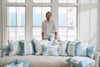 In a bright room, someone holds a light blue pillow with teal velvet welt amidst various decorative pillows, including the charming Bee & Florals Collection: Victorian Tile Pillow from Sewing Down South on a beige sofa. Natural light streams through large windows overlooking a nearby blue-paneled building.