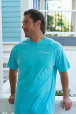 A man with medium-length hair and a beard stands on a porch, wearing a bright blue Sewing Down South Logo Rainbow Row Tee. The charming blue-and-white building and white railing in the background evoke Rainbow Row. He gazes thoughtfully to his left.