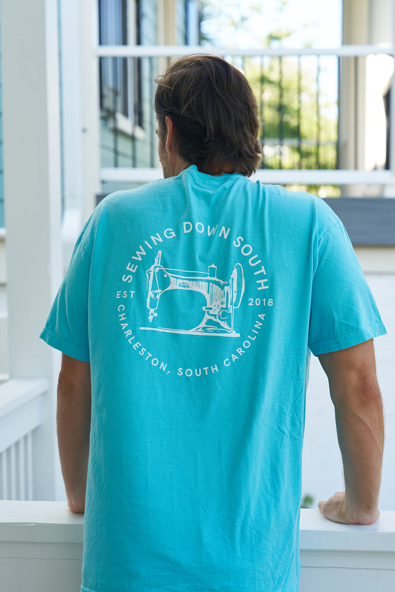 A person with brown hair stands on a porch, facing away, wearing a turquoise Comfort Colors unisex tee called the Sewing Down South Logo Rainbow Row Tee. It features a vintage sewing machine graphic and Sewing Down South, Charleston, South Carolina, Est. 2018, reflecting Rainbow Rows charm.