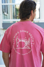 A man stands outside with blurred Rainbow Row-like buildings in the background, wearing a pink Comfort Colors T-shirt called Sewing Down South Logo Rainbow Row Tee, featuring Sewing Down South and a sewing machine graphic on the back.