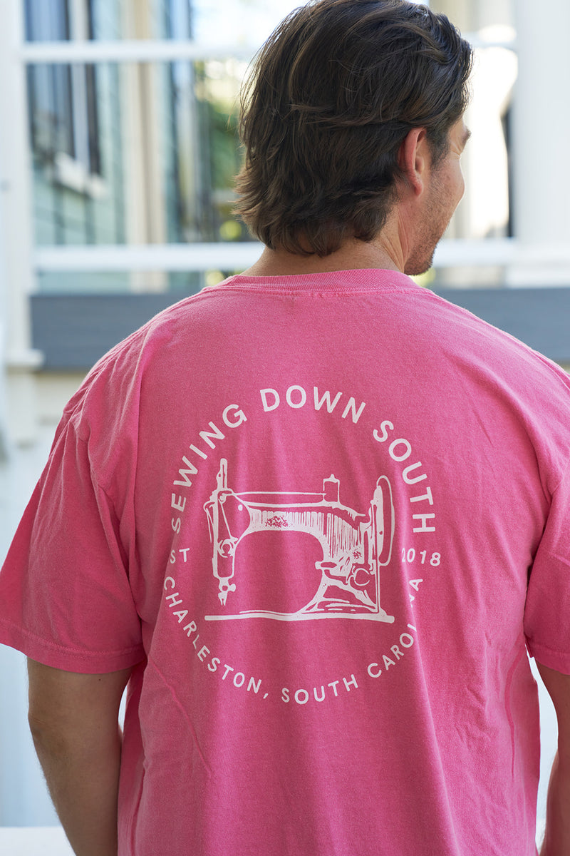A man stands outside with blurred Rainbow Row-like buildings in the background, wearing a pink Comfort Colors T-shirt called Sewing Down South Logo Rainbow Row Tee, featuring Sewing Down South and a sewing machine graphic on the back.