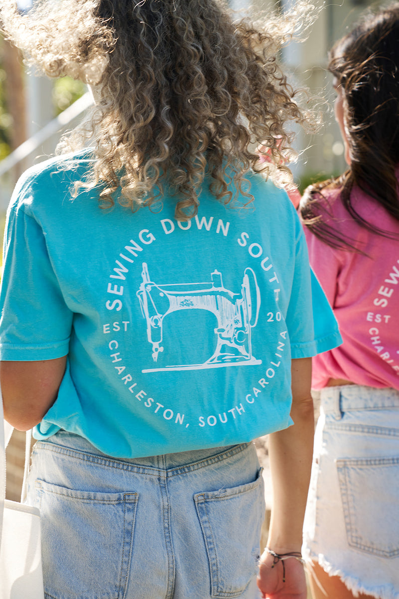 Two people stroll in vibrant unisex Sewing Down South Logo Rainbow Row Tees featuring a sewing machine logo and Sewing Down South, Est 2020, Charleston, South Carolina on the back. Made from Comfort Colors fabric, they carry a white tote, with their long hair flowing in the breeze.