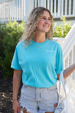 A woman with curly hair in a Sewing Down South Logo Rainbow Row Tee and denim shorts smiles by white stairs and lush greenery, holding a white bag.