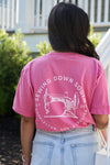 A person with long hair is wearing the pink unisex Sewing Down South Logo Rainbow Row Tee featuring a sewing machine illustration and text on the back. Designed in Comfort Colors, theyre outdoors near greenery and steps, evoking Charlestons iconic Rainbow Row charm.